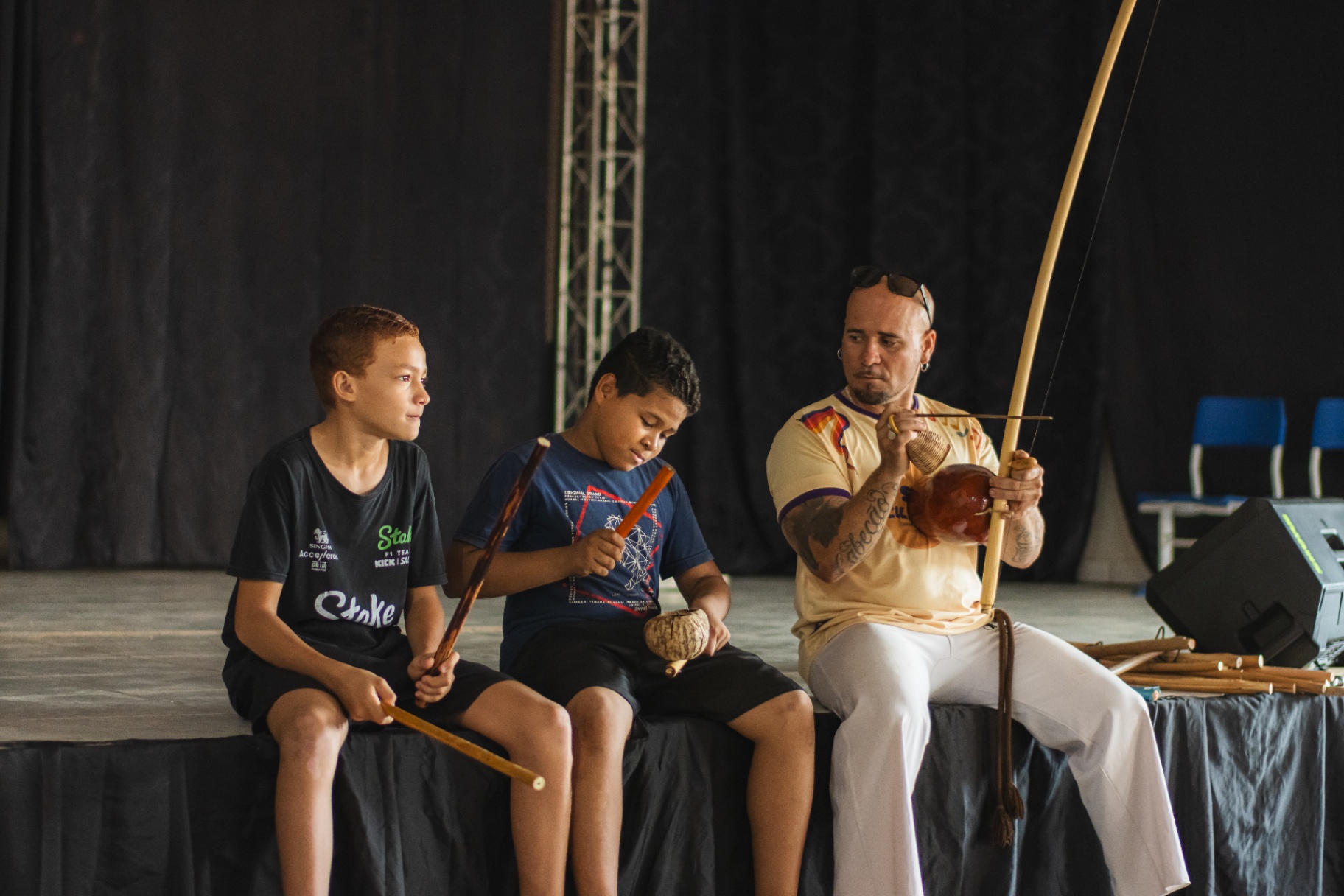 Educador ensinando dois meninos a tocar berimbau e reco-reco no palco durante o Sarau Cultural
